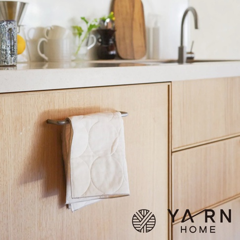 淺米色圓形絎縫廚房擦手巾，掛在淺木色櫥櫃的銀色毛巾架上。