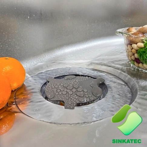 不鏽鋼廚房水槽中安裝著一個灰色的 SINKATEC 磁吸濾網蓋，濾網表面覆蓋著水珠，旁邊放置著橘子與裝有豆類食材的透明碗，水槽內有少量積水。