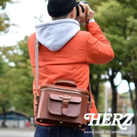 身穿橘色夾克、頭戴黑色毛帽的男子，正背著日本HERZ手工皮革相機包，在戶外使用相機拍照。