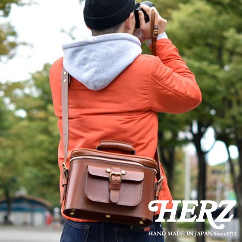 身穿橘色夾克、頭戴黑色毛帽的男子，正背著日本HERZ手工皮革相機包，在戶外使用相機拍照。