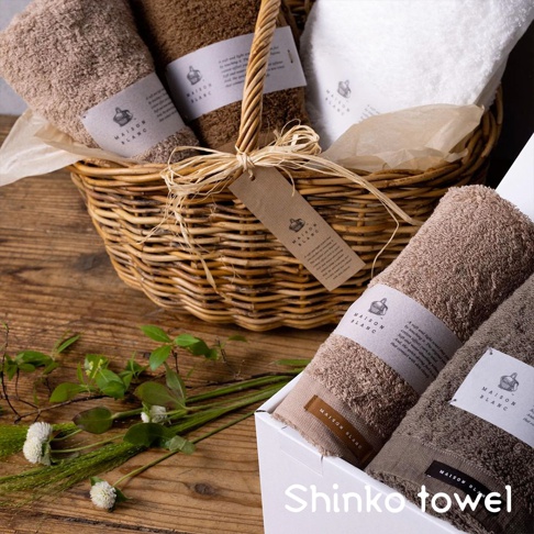 MAISON BLANC 輕奢柔膚匹馬棉毛巾，展示了咖啡色、褐色及白色毛巾，部分毛巾捲起放置於藤編籃中，並有品牌標籤。另一組毛巾擺放在白色禮盒內，旁邊有綠色植物裝飾。