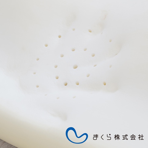 白色護頸止鼾枕，表面佈滿大小不一的透氣孔，材質呈現柔軟的記憶棉質地，中央有藍色曲線標誌與日文公司名稱「まくら株式会社」。