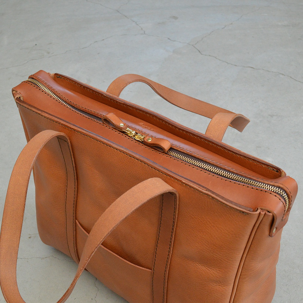 職人手作的棕褐色皮革托特包，設有拉鍊開合、兩個皮革提把，以及一個外部插袋。