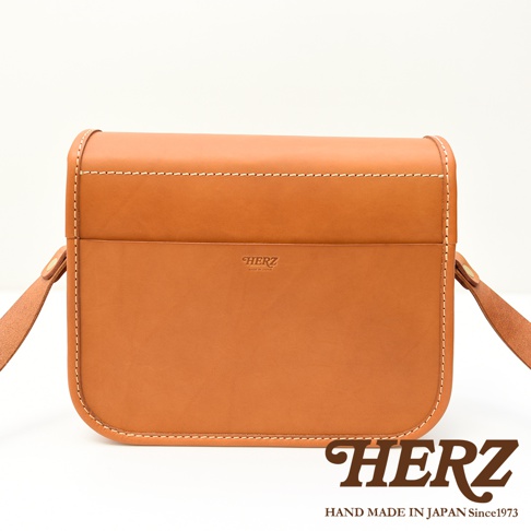 日本HERZ職人手作皮革經典雙扣郵差包，焦糖色牛皮材質，銀色雙扣設計，正面壓印HERZ品牌標誌及MADE IN JAPAN字樣，附有可調式背帶，整體呈現簡約復古風格。