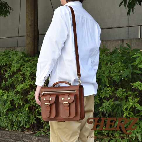 棕色皮革製的雙口袋兩用單肩包，配有頂部提把、拉鍊開合及兩個帶有銅扣的翻蓋前袋，可肩背或手提。