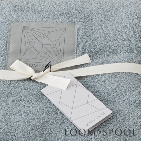 淺藍色純棉浴巾，標籤和緞帶綁成蝴蝶結，上方標籤印有幾何圖案及LOOM&SPOON字樣