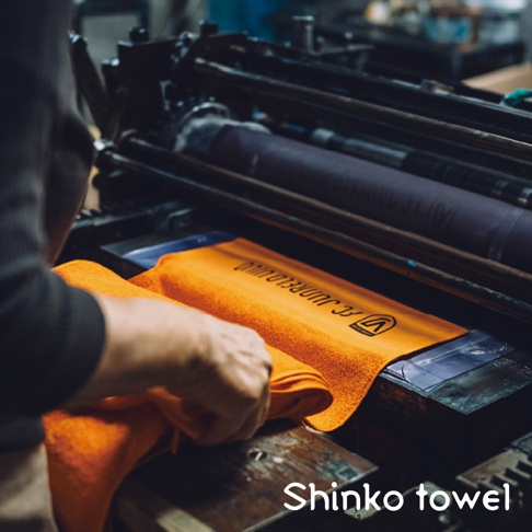 一件橘色毛巾，質地柔軟，上面印有黑色文字與 Logo，放置於印刷機台上，一隻手正在調整毛巾。