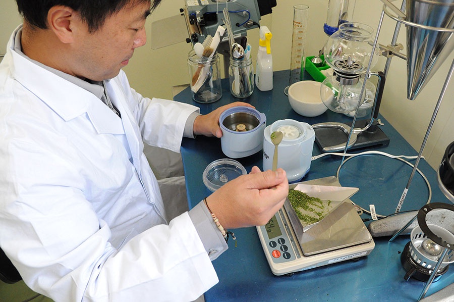 一名身穿白袍的男性正在實驗室桌前操作，桌上擺有電子秤、磨粉機、試管等實驗器材，他手持湯匙，似乎正在處理綠色粉末。