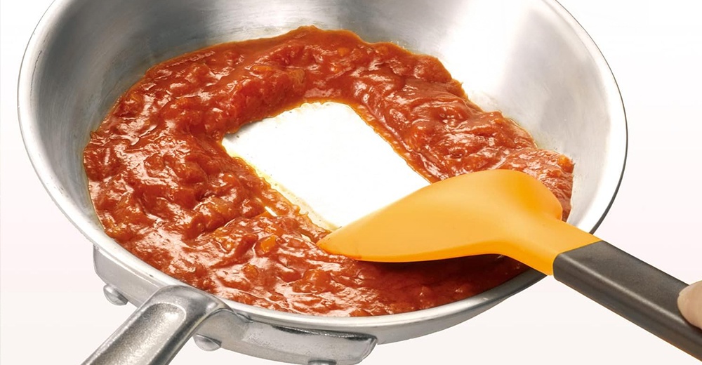 一個橘色的矽膠鍋鏟正在攪拌不鏽鋼鍋中的紅色醬料