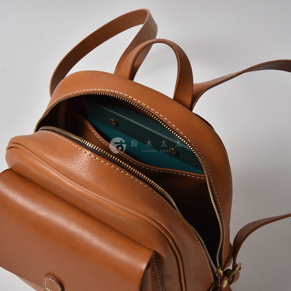 咖啡色皮革迷你後背包，設有立體前袋和拉鍊主隔層，內含綠色筆記本，搭配白色車線和金色五金配件。