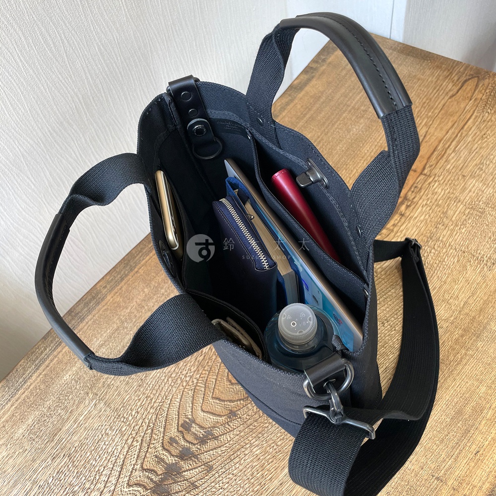 黑色帆布托特包，內部裝有手機、錢包、平板電腦、水瓶和紅色筆。包有頂部提把和可拆卸肩帶。