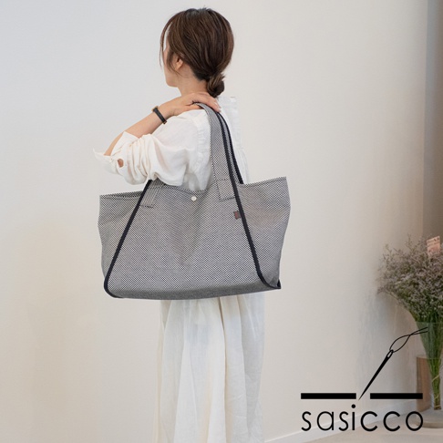 一位女士肩背一款黑色與白色細點刺子織紋理的托特包，包身寬大，有深色滾邊與提把，包口中央有鈕扣。