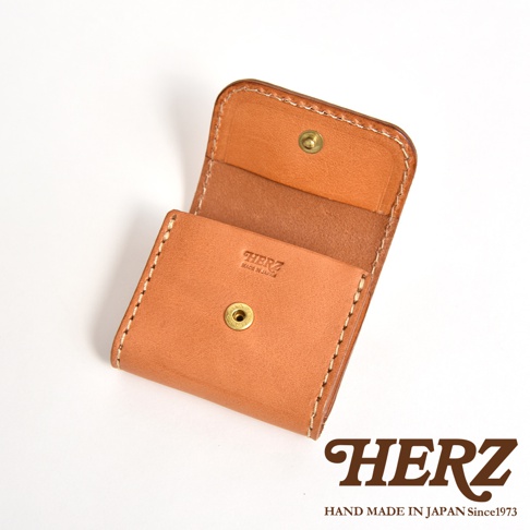 一個日本HERZ品牌職人手作的淺棕色Stare皮革雙折零錢包，展開呈現，正面印有品牌標誌與Made in Japan字樣，搭配金屬按扣設計，質感細膩且縫線工整，右下角配有品牌標誌與成立年份說明。