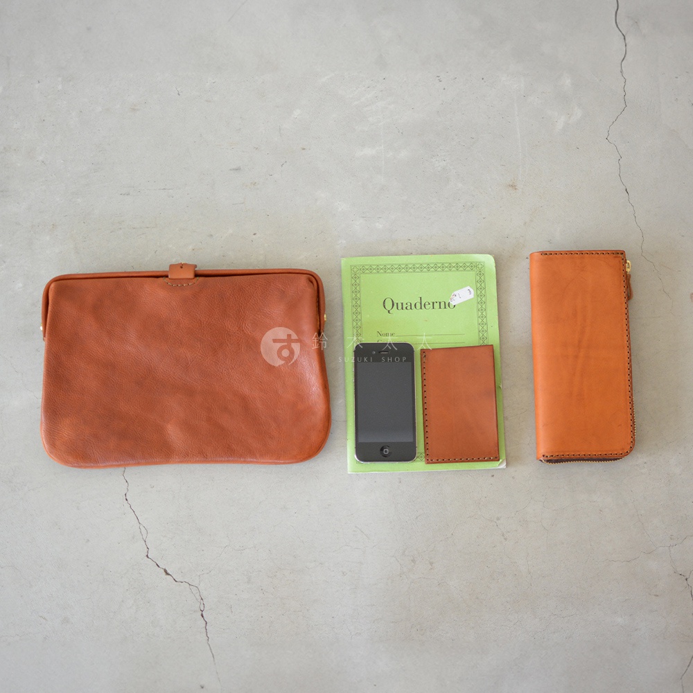 職人手作皮革Organ手拿包，呈現自然棕色皮革紋理，搭配金色金屬扣環。旁有手機、皮革證件夾及長夾，共同展示產品實用性與質感。