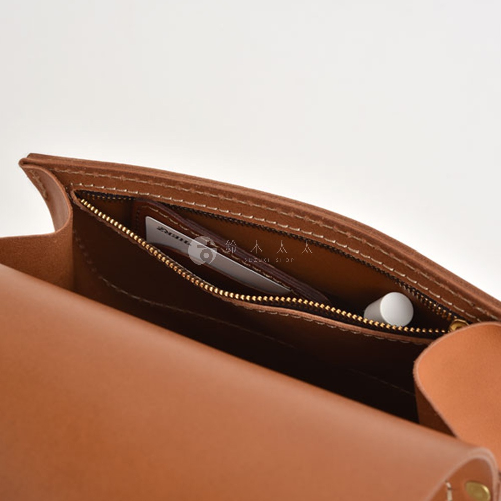 皮革翻蓋迷你後背包特寫，展示內部拉鍊夾層，內有卡片和一支白色小物，皮革為棕褐色，縫線清晰。