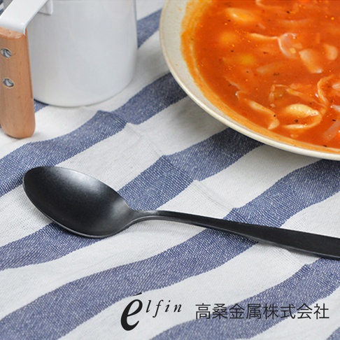 一雙日製熔岩黑石餐匙，呈現霧面黑色，材質為金屬，擺放在藍白條紋的餐巾上，一旁還有一個裝有番茄湯的白色盤子。