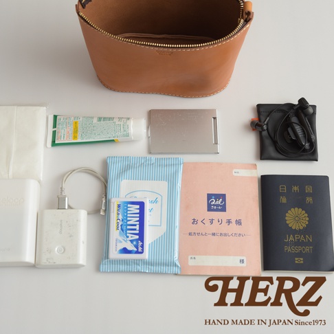 棕色皮革圓筒形收納包，內含濕紙巾、薄荷糖、手機充電器、耳機、化妝品小樣、銀色名片夾和日本護照。