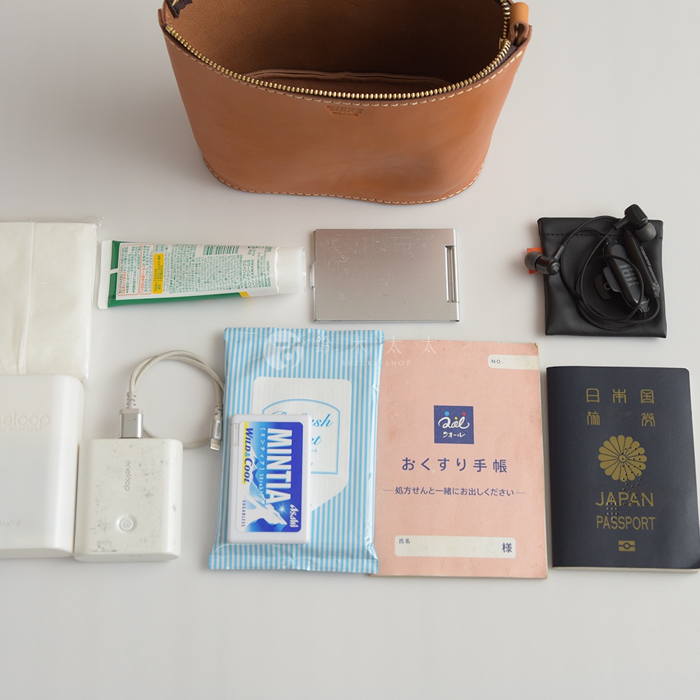 棕色皮革製成的圓筒形收納包，內有物品。下方擺放著白色面紙、牙膏、銀色鏡子、黑色耳機套、白色行動電源、濕紙巾、日文書籍和一本日本護照。