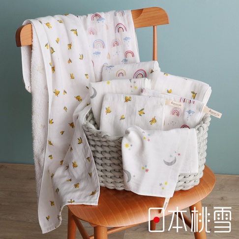 一把木椅上放著一籃子印有可愛圖案（如彩虹、檸檬、月亮和星星）的白色紗布方巾，其中一條較長的紗布毛巾懸掛在椅子扶手上。