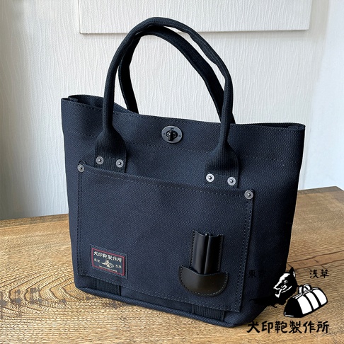 一款深藍色帆布製成的方形水桶托特包，配有兩條黑色提把，包包正面有一個帶有金屬扣的翻蓋口袋，口袋下方有一個紅色標籤，標籤上印有日文文字和圖案，旁邊還有一個黑色皮革裝飾件。