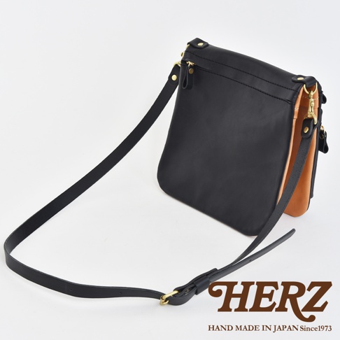 職人手作皮革翻轉雙色直式斜背包，主體為黑色皮革，側邊翻面為原色皮革，搭配金色五金配件與黑色皮革長背帶，整體風格簡約。