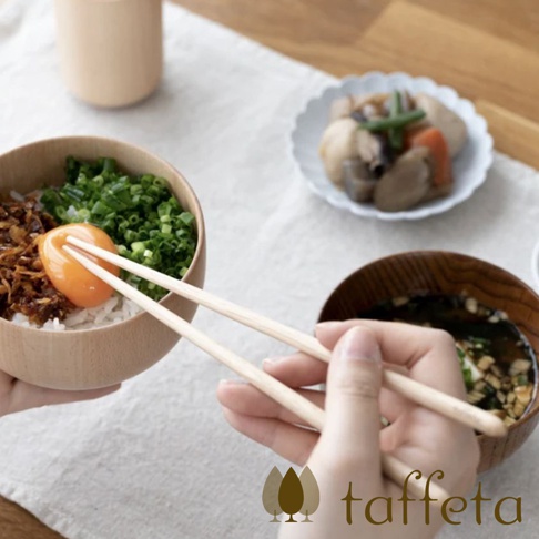 一雙23公分長的楓木圓筷，淺木色，一端夾著生雞蛋和蔥花飯，另一端伸入湯碗中。