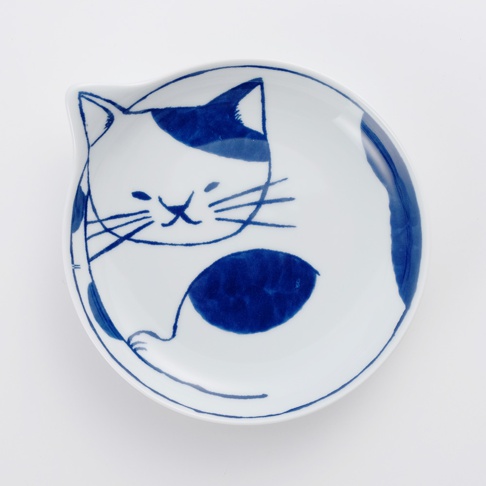 一個白底藍花的陶瓷小碟，繪有簡約的貓咪圖案，貓咪的臉部和身體呈現可愛的蜷縮姿態，藍色筆觸描繪出貓咪的耳朵、眼睛、鬍鬚、斑紋和尾巴，碟子邊緣略微上翹，造型獨特。