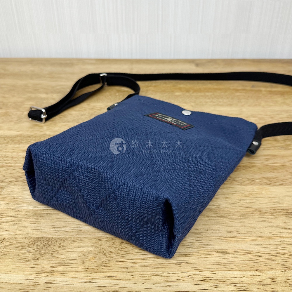 深藍色刺子織紋理平板收納側背包，配有黑色可調式背帶，包身側面有品牌標籤及金屬鈕扣。