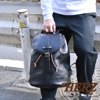 黑色皮革單扣束口後背包，具有一個主隔層，以皮革束帶和金屬扣固定，側邊有兩個可見的皮革繫繩，背包肩帶為皮革材質。