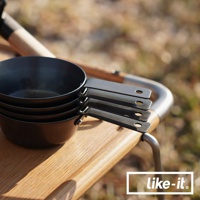 一疊黑色金屬材質的萬能調理把手鐵杯，帶有長柄，置於木製檯面上，背景模糊，顯示戶外環境。