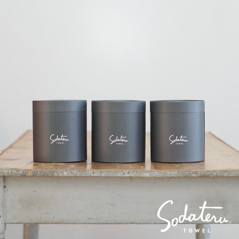 三個灰色圓筒形包裝盒，分別印有白色「Sodateru TOWEL」字樣，整齊排列在舊木桌上，背景為白色。