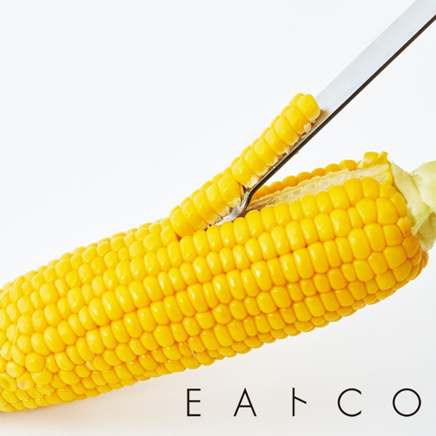 日製玉米刨刀正在刨下新鮮玉米粒，呈現飽滿的黃色，刀身為銀色金屬材質。