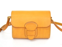 職人手作皮革寬扣帶馬鞍包，採用天然棕色皮革製成，包身結構方正，帶有精緻的縫線細節和寬版皮革扣帶，側邊有金屬掛環連接可調節的皮革背帶。