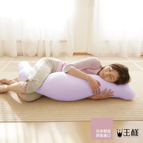 一名女子側臥在淡紫色美容抱枕上，抱枕呈彎月形，材質柔軟。女子身穿粉色上衣和灰色長褲，雙手環抱抱枕，面露舒適表情。