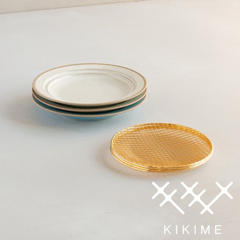 美濃燒瀝油餐盤，盤身為白色，外圈帶有淺棕色邊緣，下方堆疊著藍綠色盤子。一旁擺放著金色圓形網架，網架表面為細密格紋。