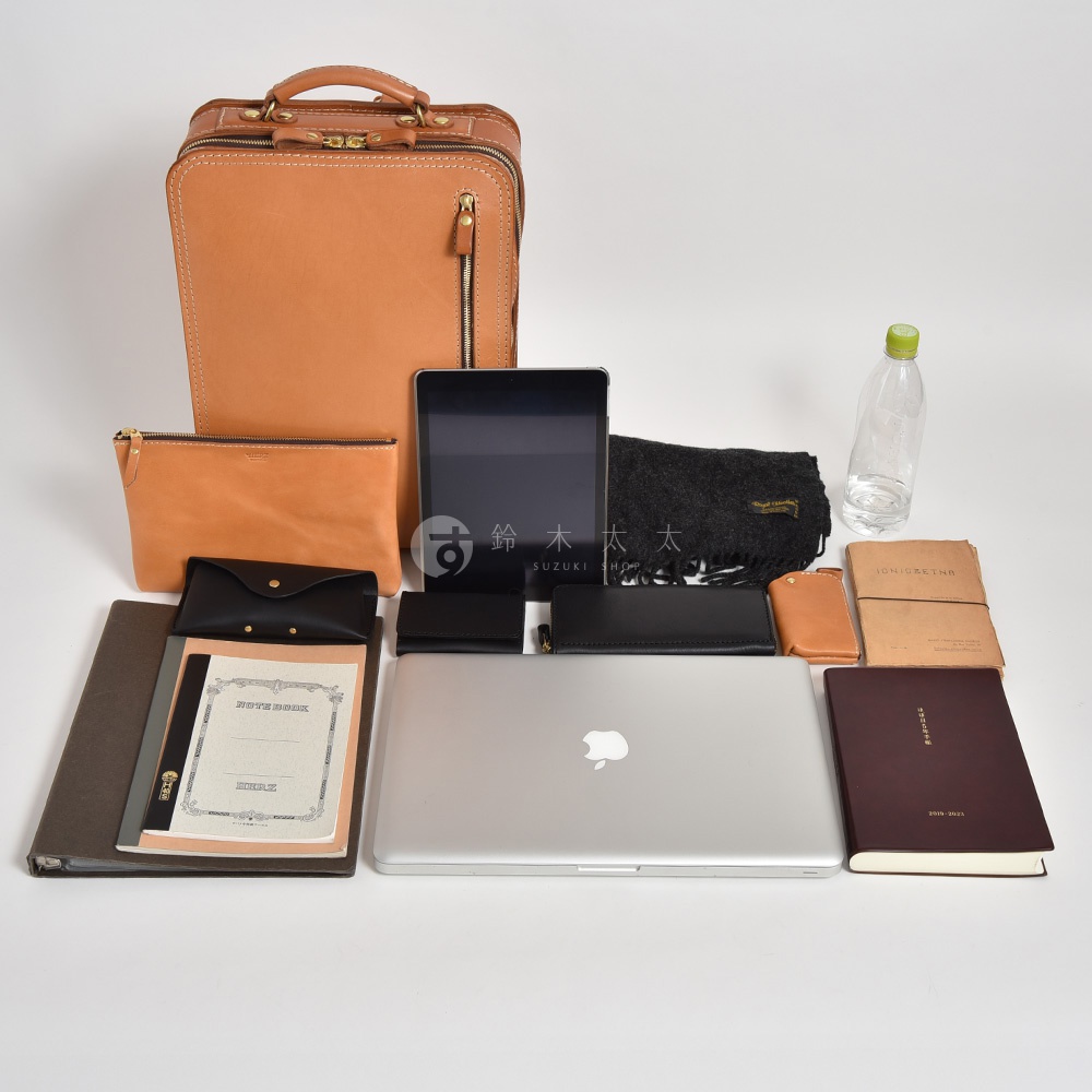 淺棕色皮革製成的手提電腦後背包，搭配拉鍊開口。側邊有一個淺棕色皮革手拿包，上方有一個黑色皮革零錢包。背包前有一台銀色筆記型電腦，上方有一個黑色平板電腦。右側有黑色圍巾、一個淺棕色卡夾、一本米色筆記本和一本深紅色記事本。