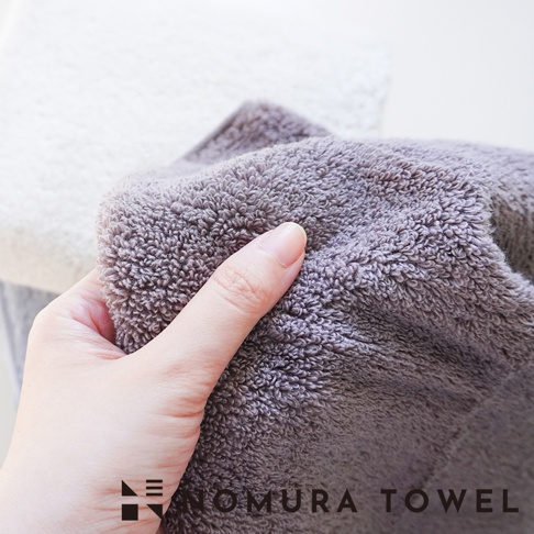 野村毛巾 YABAI 水漾瞬吸厲害毛巾，手觸灰色毛巾，上面有 NOMURA TOWEL 字樣。