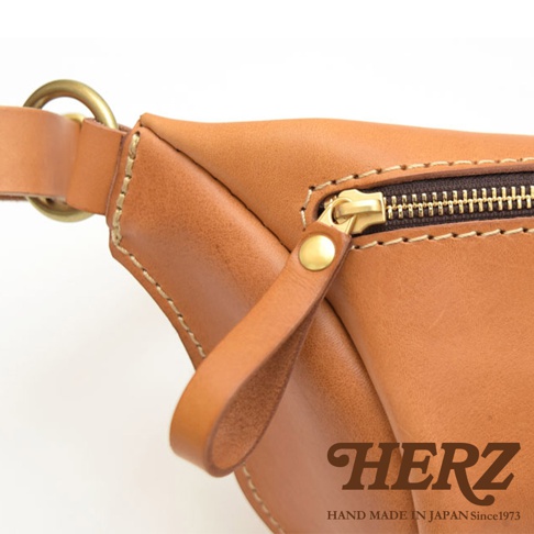 職人手作的棕色皮革輕便斜背包，特寫拉鍊、皮革拉環和黃銅色金屬扣環，縫線清晰可見。