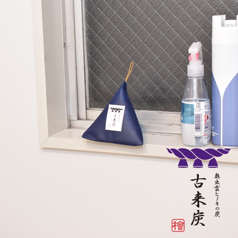 一個深藍色的立體三角型調濕木炭產品，表面有細緻的紋理，頂部有黃色細繩。產品上貼有白色標籤，標籤上有日文文字和黑色圖案。產品擺放在窗台，旁邊還有一個透明噴霧瓶和一個藍白色噴霧罐。
