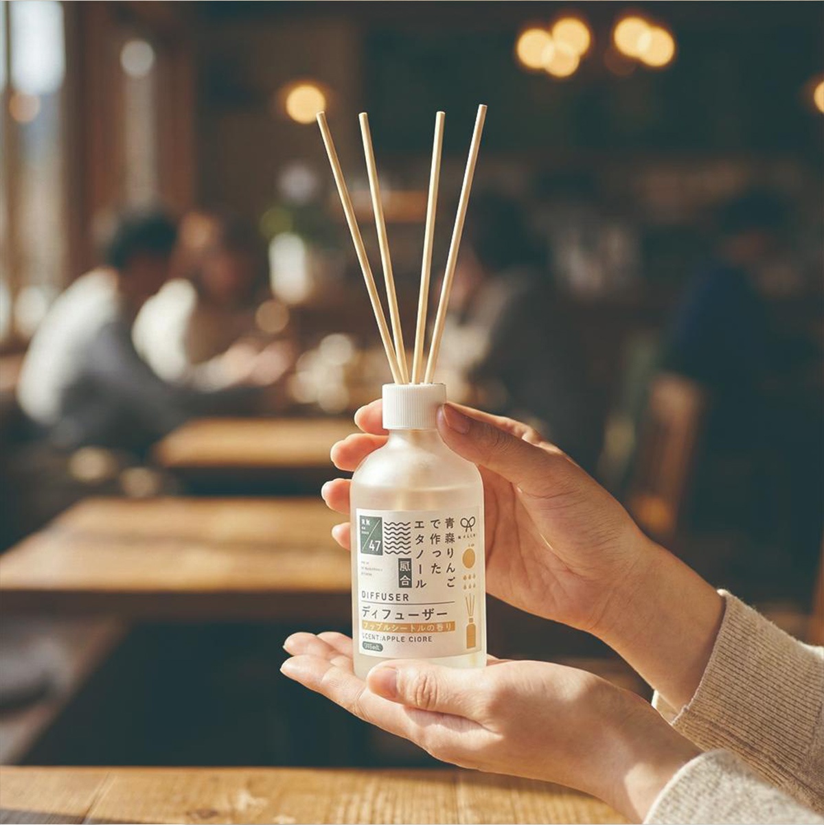 一雙手捧著裝有擴香竹的日式室內擴香瓶，瓶身貼有日文標籤，背景是模糊的咖啡館或餐廳場景。