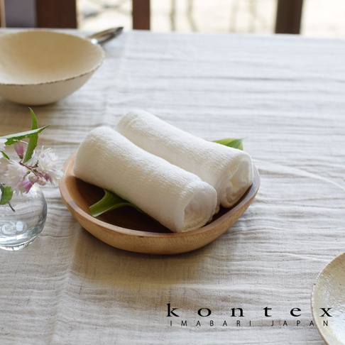 兩條白色紗布巾捲起，放置於淺木碗中，碗底鋪有綠葉。背景為白色桌布，左側有玻璃花瓶裝飾著粉色小花。