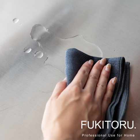 一隻手正拿著深灰色超細纖維擦拭布，擦拭佈滿水滴的銀色金屬檯面。