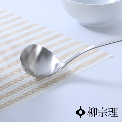 柳宗理不鏽鋼湯勺放置在條紋餐墊上，湯勺設計簡約且具有圓潤的弧度，背景角落設有一只白色瓷碗，右下方印有品牌名稱柳宗理。