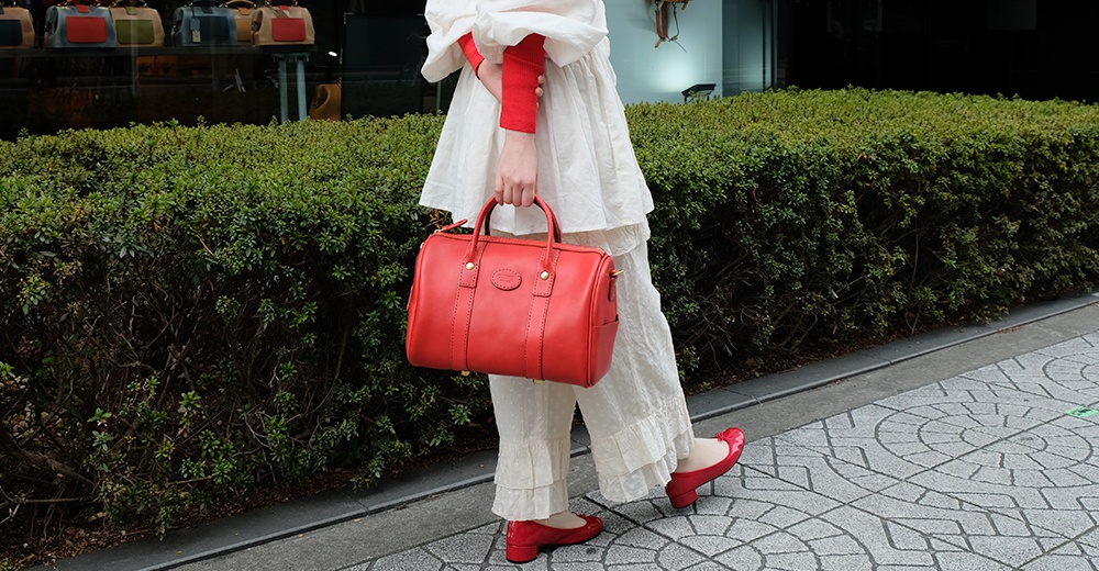 紅色皮革波士頓包，有手提帶和可拆卸肩帶，頂部有拉鍊開合，正面有品牌標誌和對稱縫線細節。