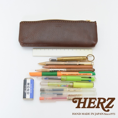 職人手作皮革經典收納筆袋，深棕色皮革材質，帶有金色拉鍊。筆袋下方擺放著一支黃銅子彈造型筆、一支金色自動鉛筆、兩支鉛筆、一支橘色筆、一支綠色筆、兩支透明筆桿的筆，以及一塊藍色擦拭布。
