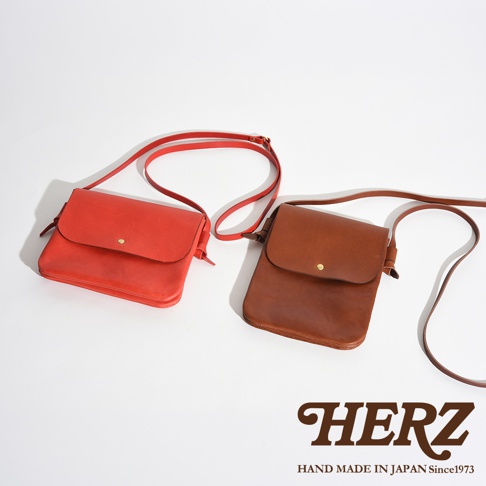 這張照片展示了兩個日本 HERZ 品牌的手工皮革隨行袋，左側為紅色的橫式包款，右側為棕色的直式包款，兩者均配有細長背帶與簡約的金屬釦環，品牌商標印於右下方。