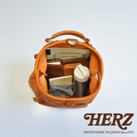 職人手作皮革輕旅行後背包 R-144，展示為橘棕色皮革，包內裝有筆記本、錢包、水瓶及毛巾等物品，包款呈現圓潤造型，配有提把及金屬扣環。