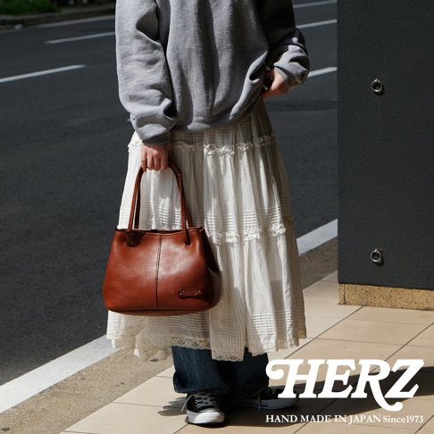 HERZ 職人手作棕色皮革托特包，搭配白色蕾絲長裙，左側為簡潔的灰色牆面。