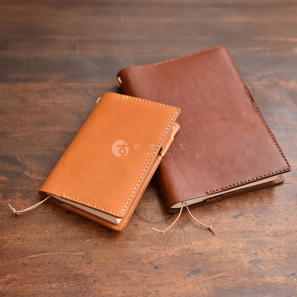 兩個皮質手帳書套，一個為淺棕色，一個為深棕色，擺放在木紋桌面上。