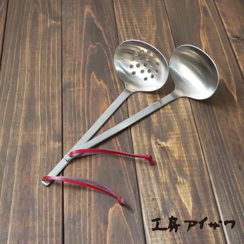 兩支不鏽鋼湯勺，一支帶有圓孔，另一支實心，均配有紅色皮革掛繩，放置在深棕色木紋桌面上。